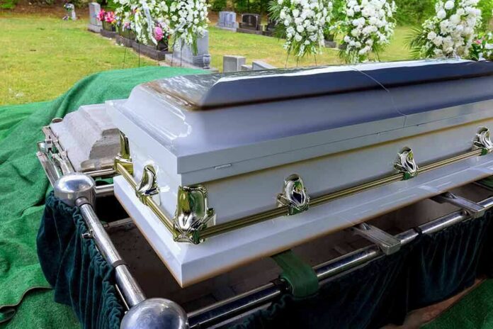 2414423041 featured image White coffin at a cemetery with flowers nearby.