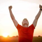 Man in a red shirt celebrating with arms raised against a sunset