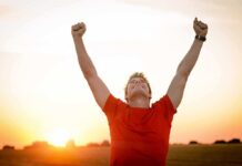Man in a red shirt celebrating with arms raised against a sunset