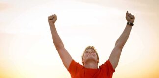 Man in a red shirt celebrating with arms raised against a sunset