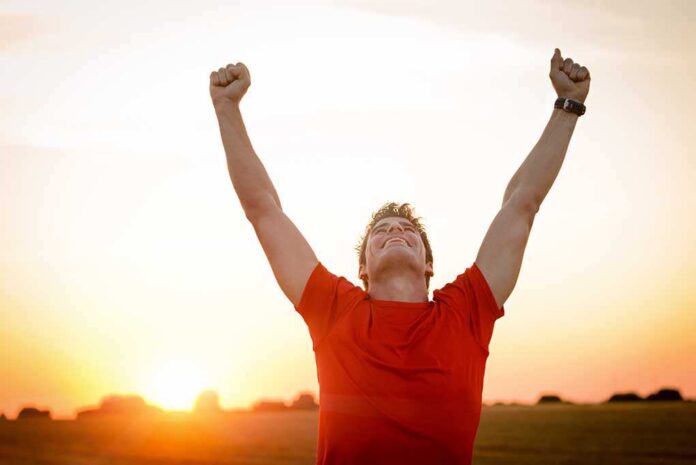 Man in a red shirt celebrating with arms raised against a sunset
