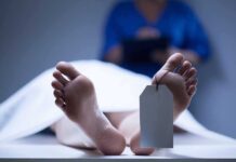 A deceased body on a table with a tag on the foot, and a medical professional in the background