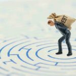 A miniature businessman carrying a sack labeled 'DEBT' in a maze