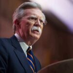 Classified Documents Found – Bolton Indicted A man in a suit speaking at a podium during a conference