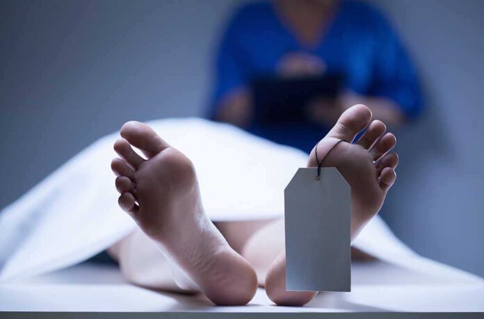 A deceased body on a table with a tag on the foot, and a medical professional in the background