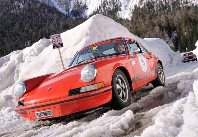 A classic orange Porsche car driving through a snowy landscape during a winter rally