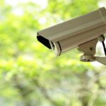 Massive Camera Hack Exposes 120K Homes A security camera mounted outdoors with a blurred green background