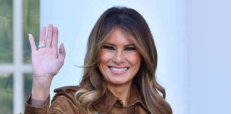 A woman smiling and waving while wearing a brown coat
