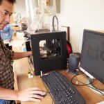 A person working at a desk with a 3D printer and computer
