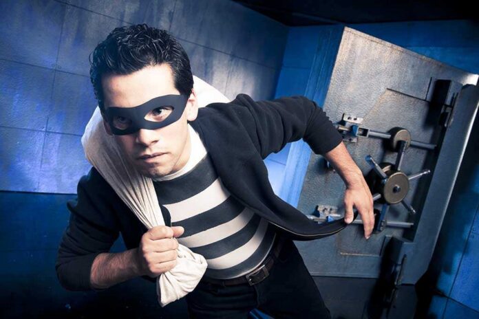 A masked thief in a striped shirt holding a bag while emerging from a bank vault