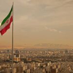 DEVASTATING Blow Leaves Iran Defenseless Against Trump Iranian flag waving over a city skyline with mountains in the background