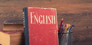 A vintage English book leaning against a stack of books next to a pen holder filled with writing instruments
