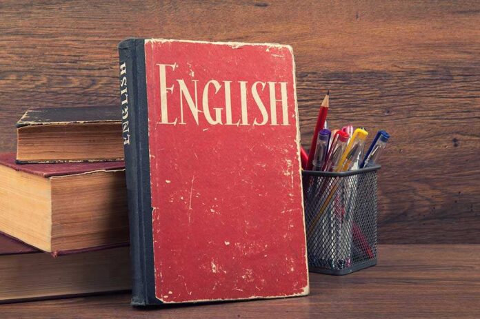 A vintage English book leaning against a stack of books next to a pen holder filled with writing instruments