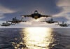Five fighter jets flying over the ocean at sunset.