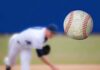 A baseball in mid-air with a pitcher in the background preparing to throw