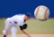 A baseball in mid-air with a pitcher in the background preparing to throw