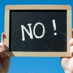 A hand holding a chalkboard with the word NO written on it against a blue sky