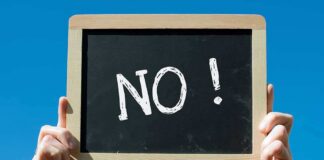 A hand holding a chalkboard with the word NO written on it against a blue sky