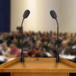 A wooden podium with two microphones in front of a blurred audience