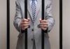 Businessman in a suit holding prison bars with handcuffs on his wrists