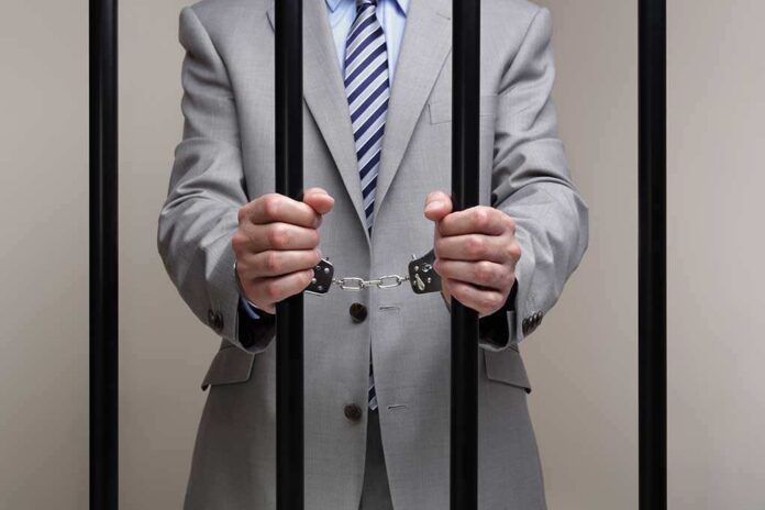 Businessman in a suit holding prison bars with handcuffs on his wrists