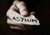 A hand holding a torn piece of paper with the word ASYLUM written on it