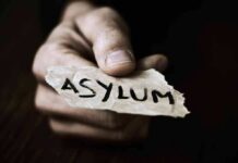 A hand holding a torn piece of paper with the word ASYLUM written on it