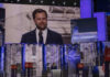 Man speaking at podium on stage, projection behind him.