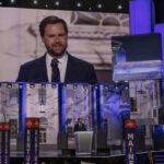 Man speaking at podium on stage, projection behind him.