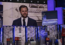 Man speaking at podium on stage, projection behind him.