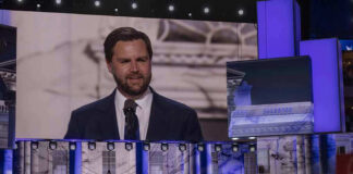 Man speaking at podium on stage, projection behind him.