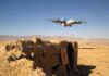 Military aircraft flying over a desert with an abandoned bunker and a for sale sign