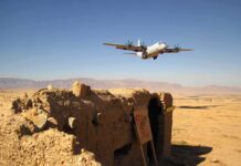 Military aircraft flying over a desert with an abandoned bunker and a for sale sign