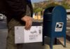 Person mailing a ballot envelope at a blue USPS street mailbox