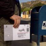 Person mailing a ballot envelope at a blue USPS street mailbox