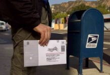 Person mailing a ballot envelope at a blue USPS street mailbox