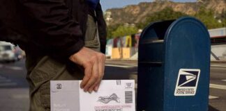 Person mailing a ballot envelope at a blue USPS street mailbox