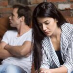 woman looking down while man sits with arms crossed on couch
