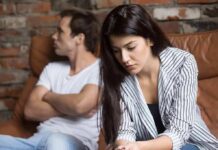 woman looking down while man sits with arms crossed on couch
