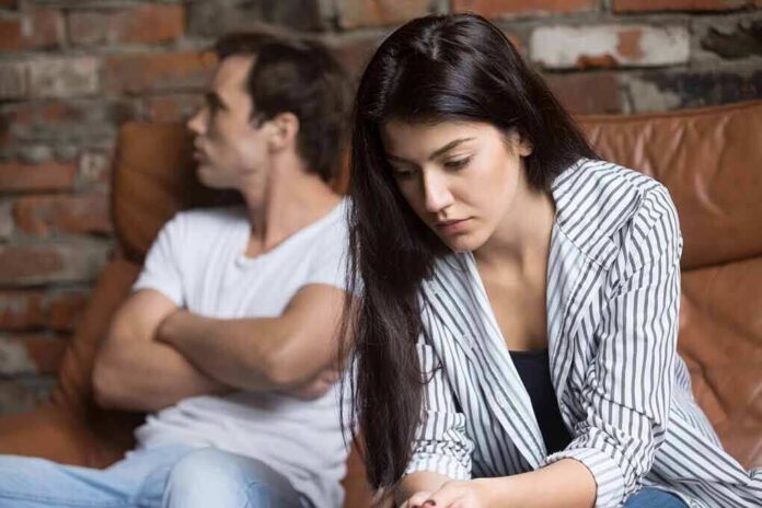 woman looking down while man sits with arms crossed on couch