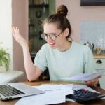 Young woman with glasses frustrated at laptop while holding bills
