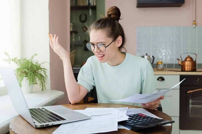 shutterstock_1119919928 Young woman with glasses frustrated at laptop while holding bills