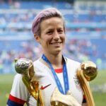 Megan Rapinoe holding soccer trophies on a stadium field