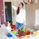 Mother standing in messy living room with toddler on couch