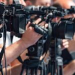 Safety Claims, Displacement Fears television cameras on tripods at a media event
